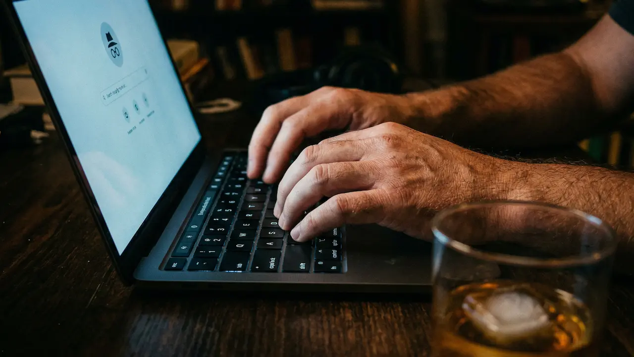Mains sur un clavier d'ordinateur dans l'obscurité, verre de whisky en arrière-plan